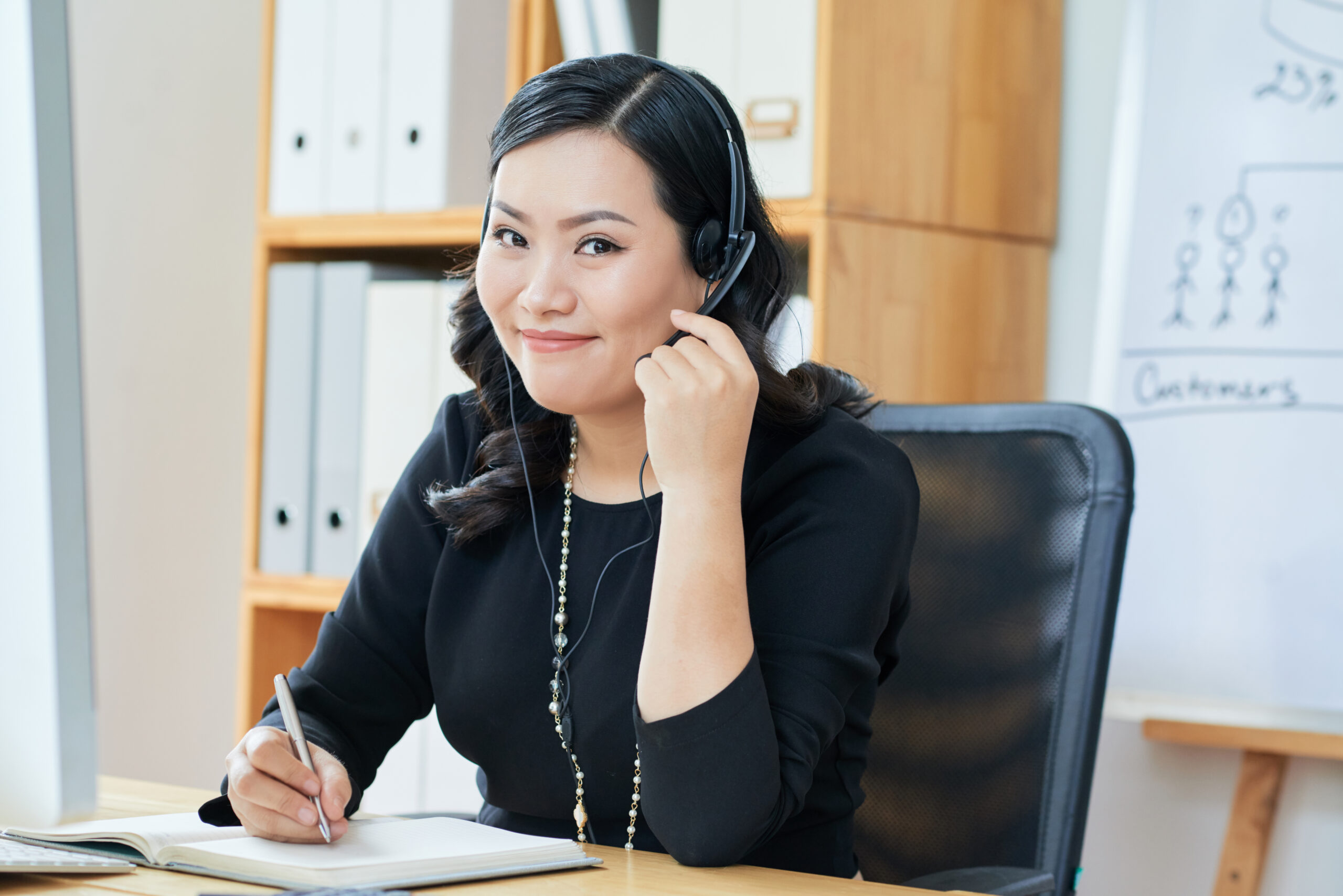 Portrait of joyful funny Asian businesswoman in headset writing in her planner